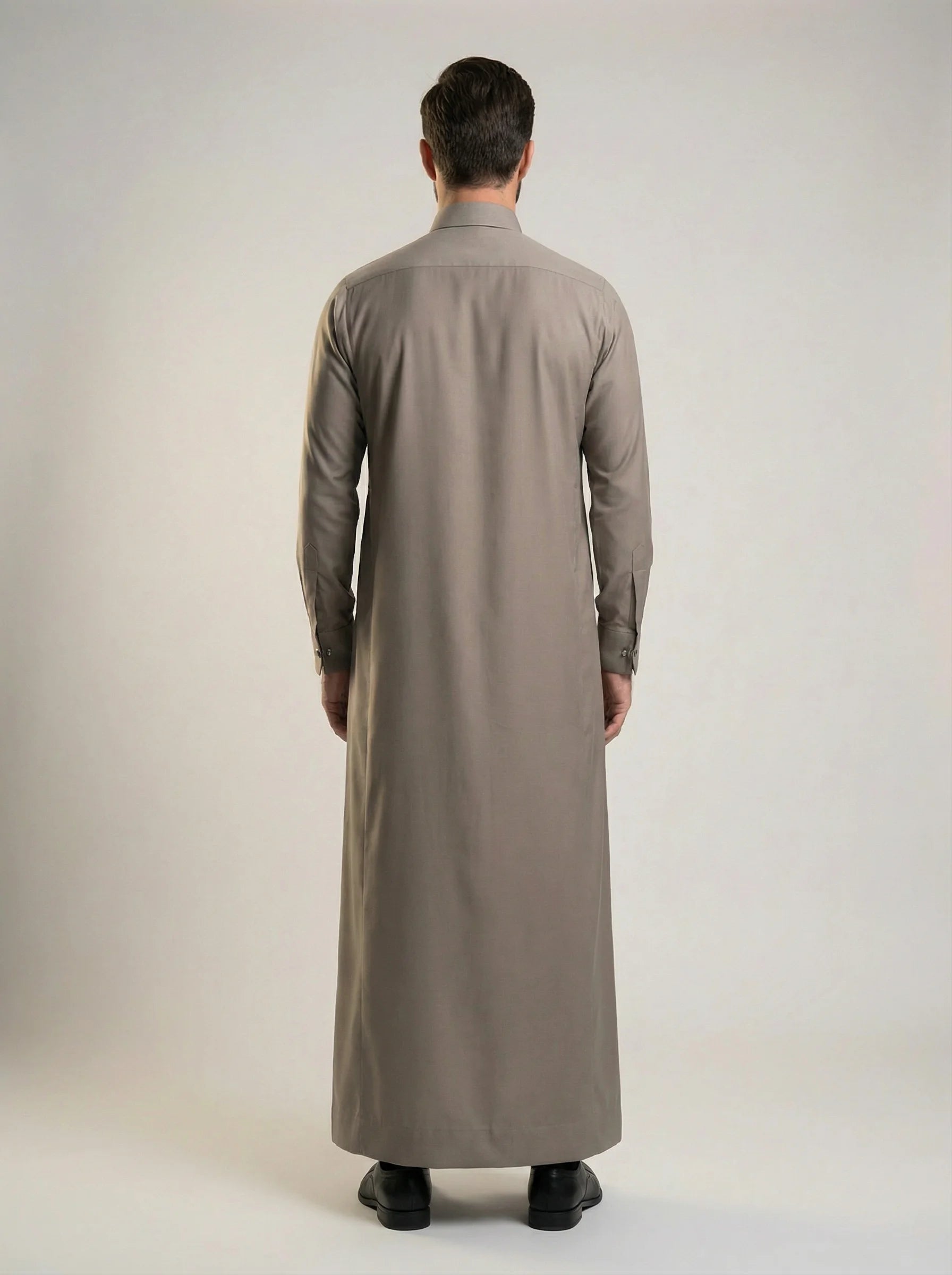 Back view of Warm Grey Luxury Thobe highlighting its clean tailored construction, graceful fabric drape, balanced silhouette, premium craftsmanship, and elegant ceremonial styling crafted for weddings, formal occasions, and refined Gulf fashion. منظر خلفي لثوب رمادي دافئ فاخر يظهر القصة المتوازنة والانسيابية الأنيقة والتفصيل الاحترافي المناسب للمناسبات الراقية والاحتفالات الرسمية.